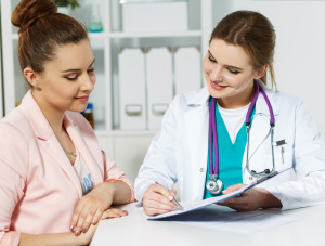 Beautiful smiling female medicine doctor holding clipped pad with document and pointing in it with pen to patient. Signing treatment bill or insurance policy concept